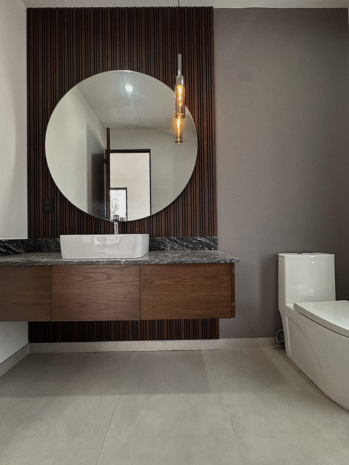 Baño de visitas con lambrín de madera y espejo circular · Casa Britania en Morelia